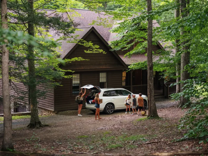 White SUV parked next to a wooden cabin in a forest, with people unloading gear.