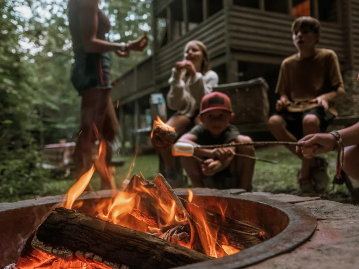 Campfire burning in a fire pit with people sitting around it near a wooden cabin in the woods.