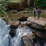 Two people standing on a large rock in a river. Small waterfalls cascade over rocky ledges on both sides with forest vegetation surrounding the scene.