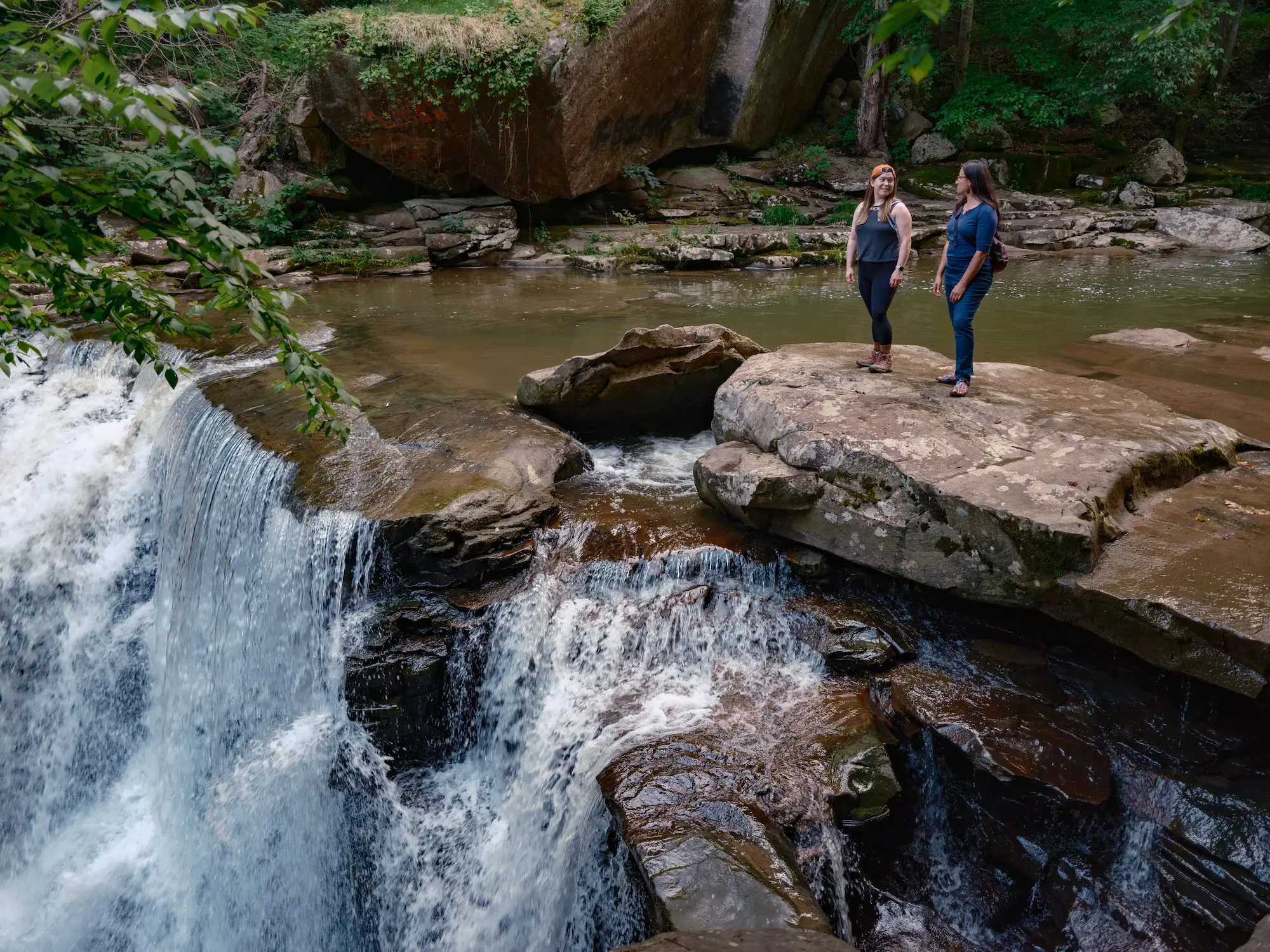 Two people standing on a large rock in a river. Small waterfalls cascade over rocky ledges on both sides with forest vegetation surrounding the scene.
