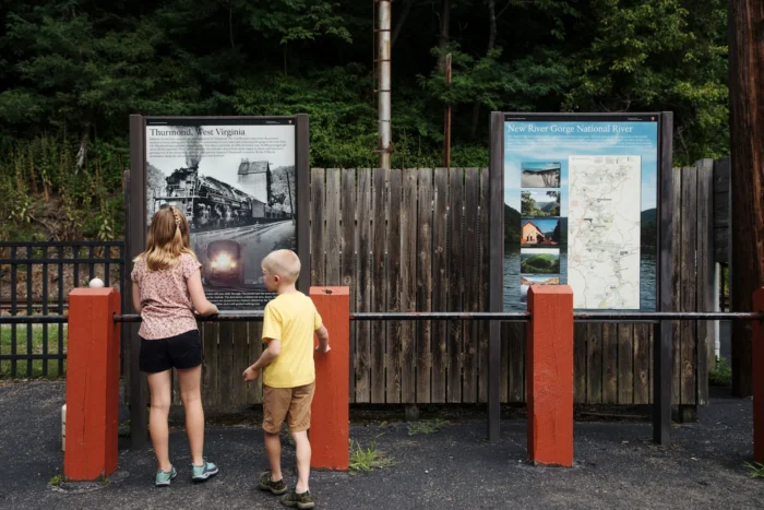 Two children reading informational signs about Thurmond, West Virginia and New River Gorge National River at an outdoor display with a wooden fence and forest background.