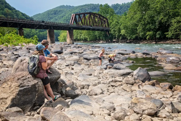 A family sitting on rocks by a river with a railroad bridge in the background and forested hills.