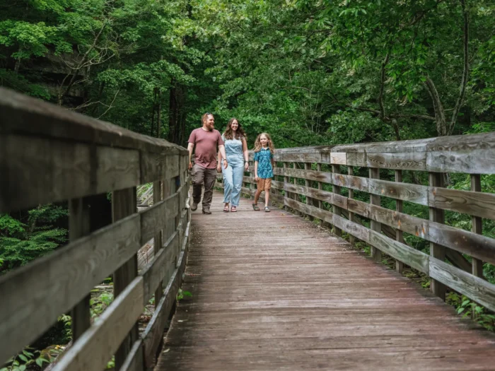 A family of three walking together on a wooden bridge surrounded by green forest.