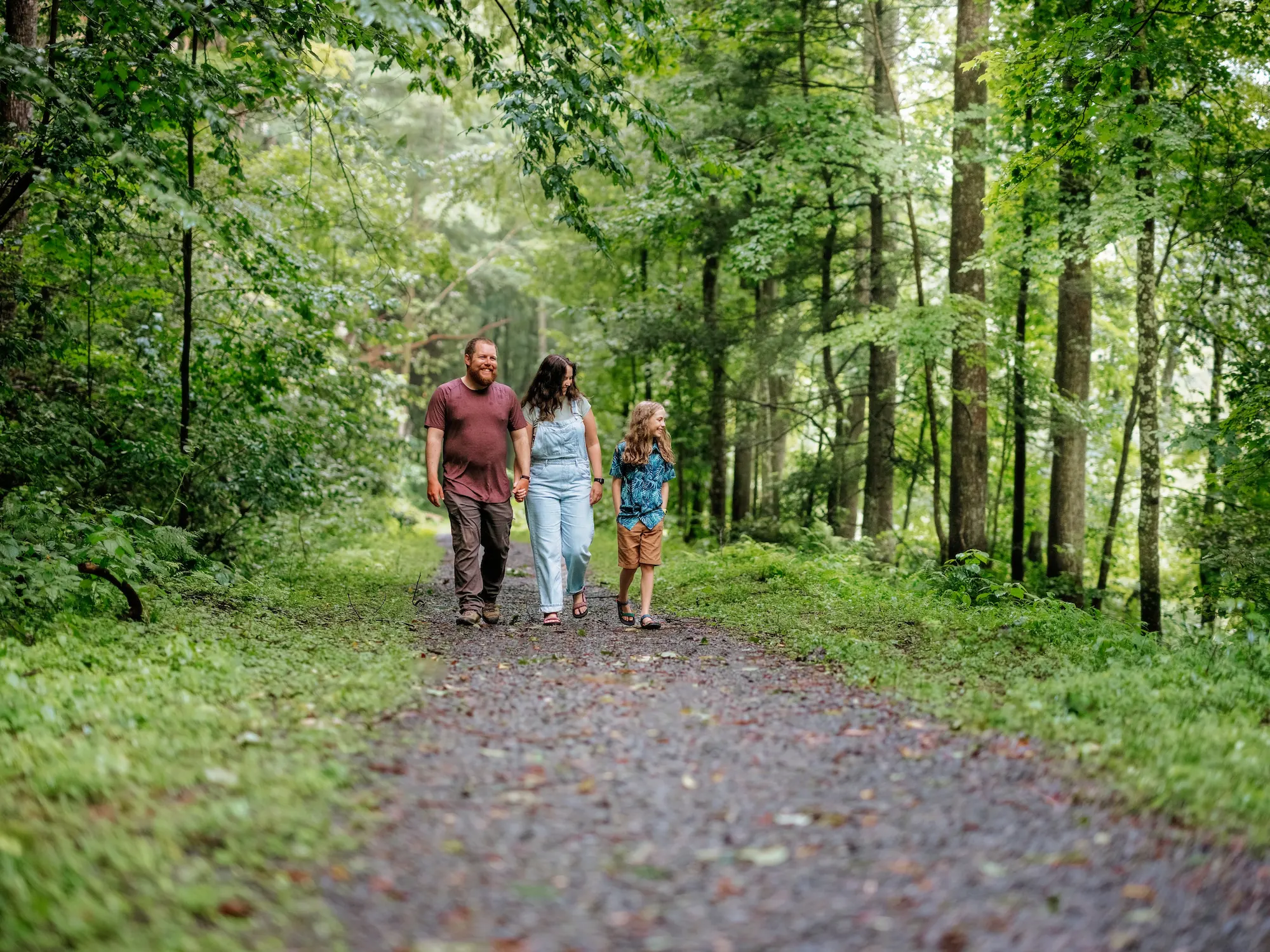 A family of three walking on a gravel path through a lush green forest.