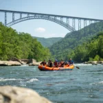 Orange raft with people white water rafting on a river, with the New River Gorge Bridge in the background.