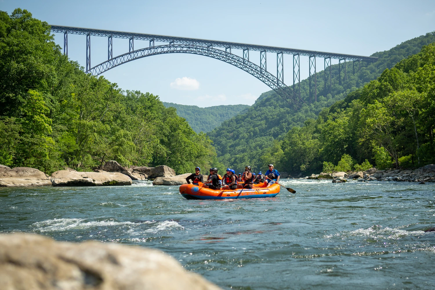Orange raft with people white water rafting on a river, with the New River Gorge Bridge in the background.