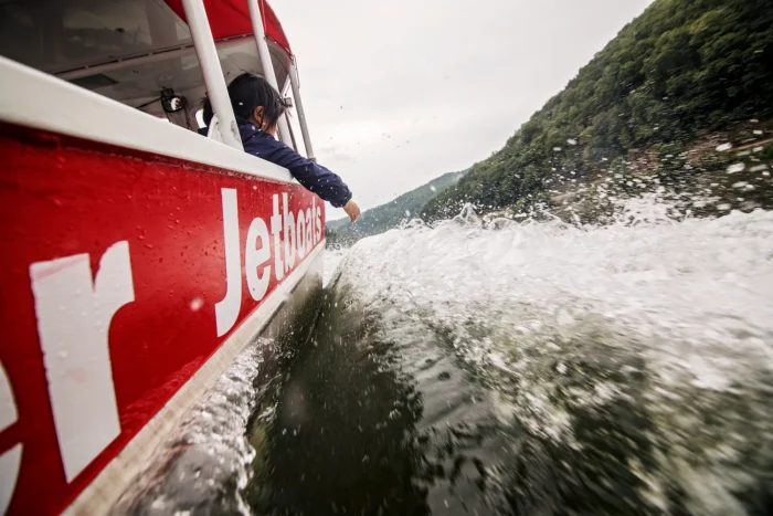 Person on a red and white jet boat traveling at high speed through river rapids, creating water spray.