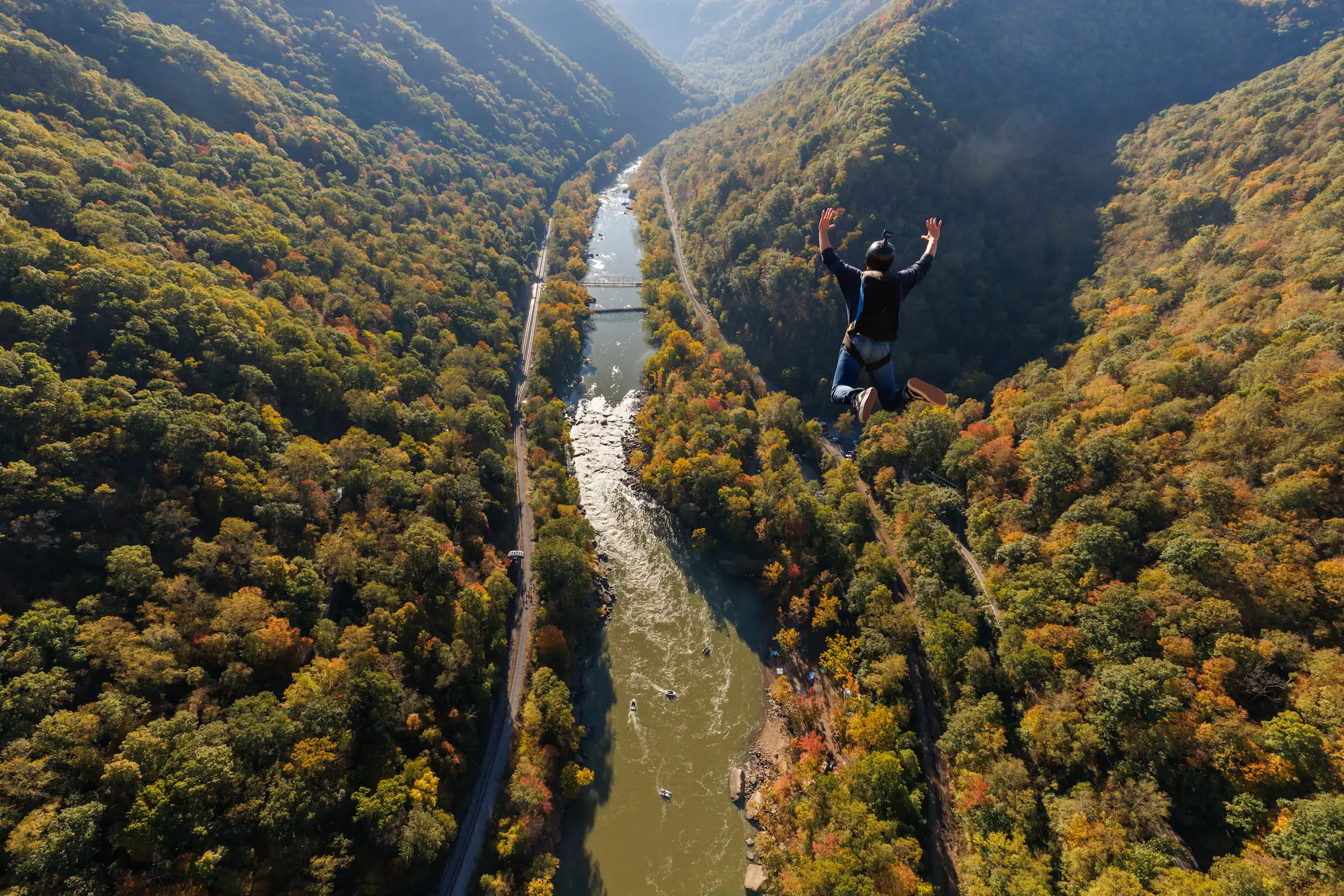 Person jumping with arms raised above the New River surrounded by autumn foliage, with a bridge visible below