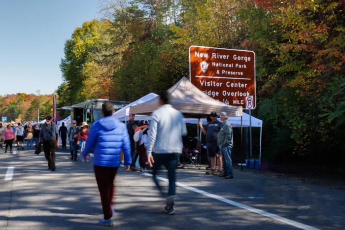 Crowd of people walking past vendor tents near a New River Gorge National Park sign during autumn with colorful fall foliage
