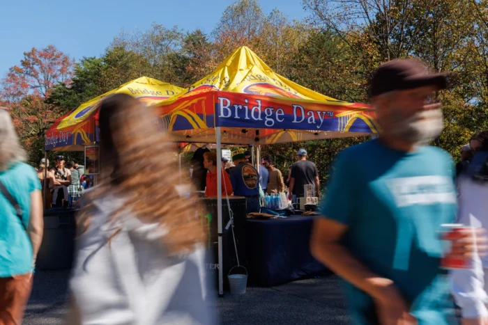 Motion-blurred people walking past a yellow and red Bridge Day event tent with autumn trees in the background
