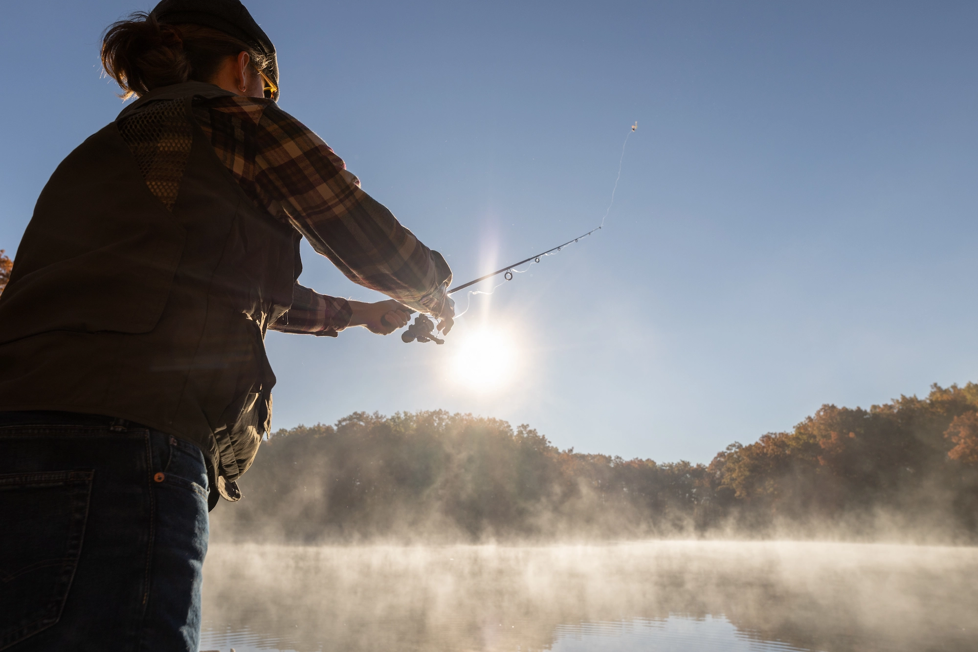 Person fishing from shore at sunrise, silhouetted against a misty lake with forested hills in background.
