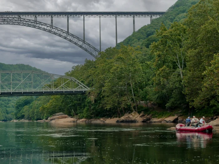 People in a red raft on a calm river with two steel arch bridges spanning across a forested valley under a cloudy sky.