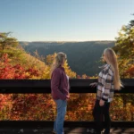 Two women viewing the New River Gorge Bridge from overlook surrounded by fall foliage
