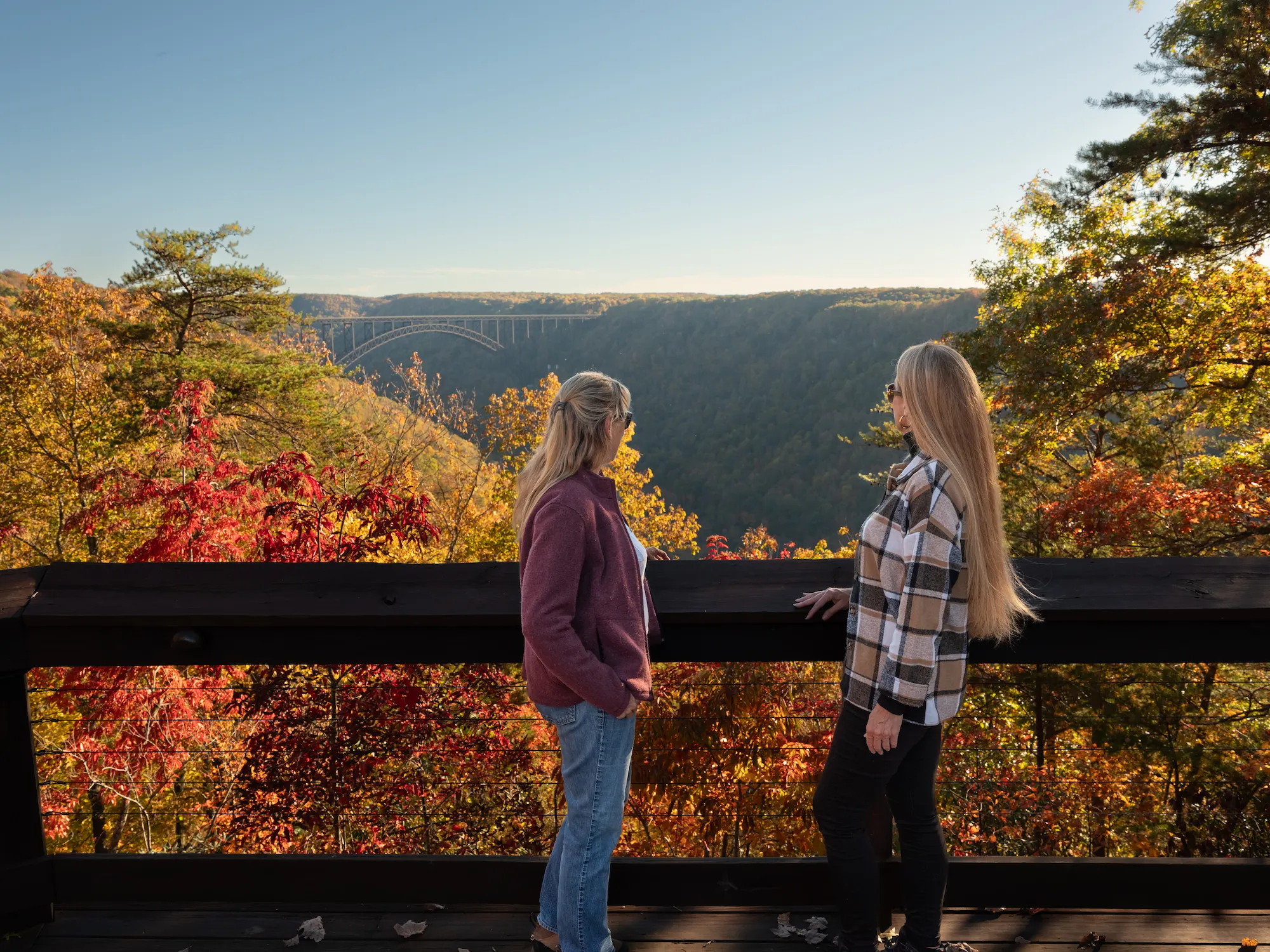 Two women viewing the New River Gorge Bridge from overlook surrounded by fall foliage