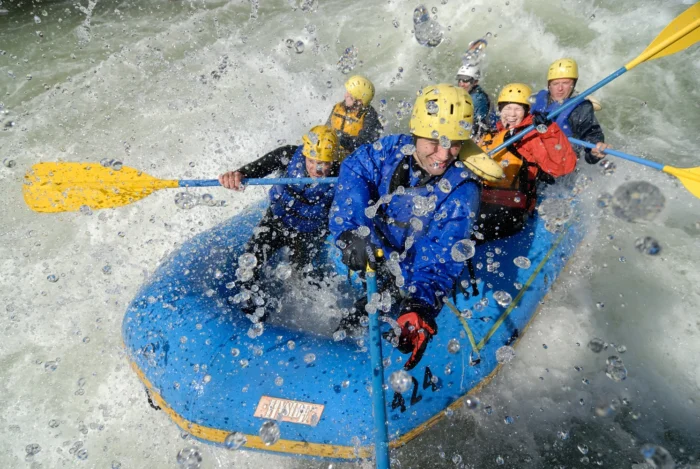 Rafters in blue inflatable raft getting drenched by rapids spray, wearing yellow helmets and life jackets while paddling with yellow oars through churning whitewater.