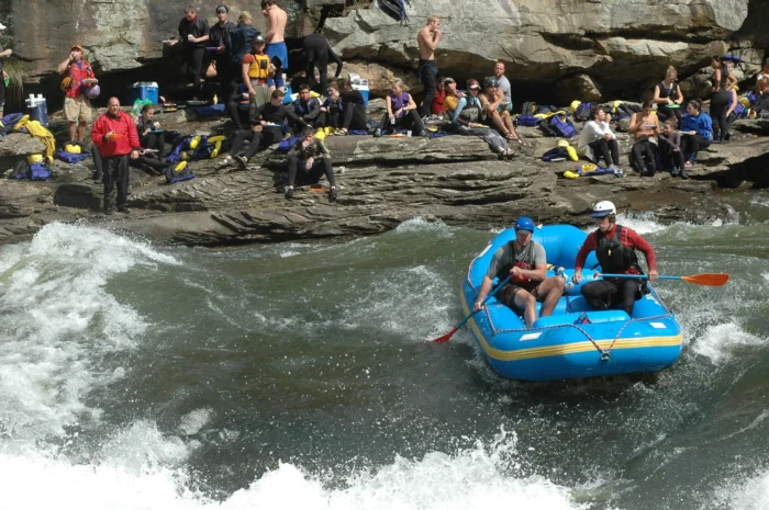 Small blue raft with three people navigating river rapids while large group of rafters and spectators sit on rocky riverbank watching, with rafting gear and equipment scattered on the rocks.