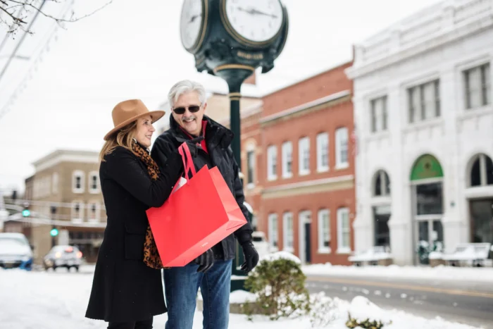 A couple shopping together in winter, holding red shopping bags while standing near a clock post in the snowy downtown Fayetteville area.