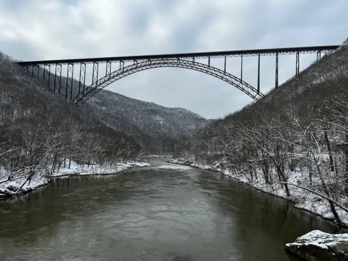 The New River Gorge Bridge spanning across the New River with snow-covered mountains and bare trees on both banks under an overcast sky.
