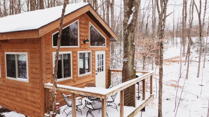 Modern wooden treehouse cabin with white-trimmed windows and snow-covered deck nestled in winter woods
