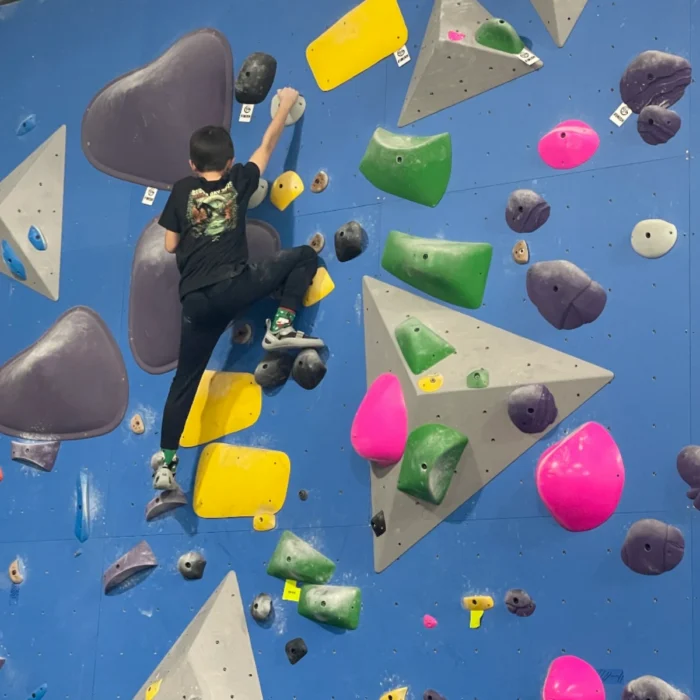 A young climber works their way up a blue indoor bouldering wall covered in colorful holds