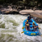Four rafters and a guide in a blue raft paddling through a rapid on the New River, with large flat rocks in the background.