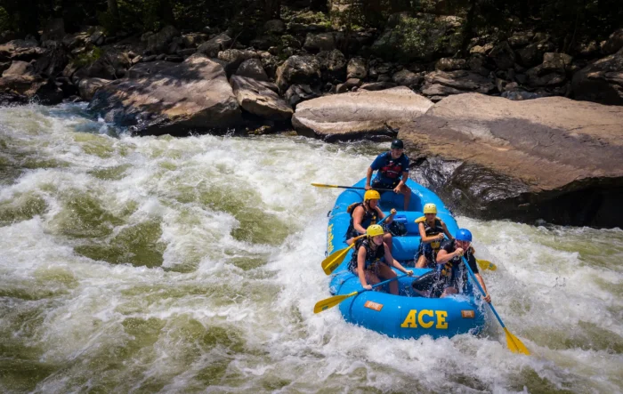 Four rafters and a guide in a blue raft paddling through a rapid on the New River, with large flat rocks in the background.