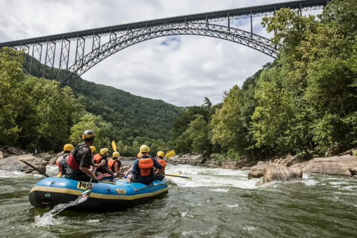 Rafters in a blue raft paddle toward the New River Gorge Bridge, framed by green trees and cloudy blue sky. 