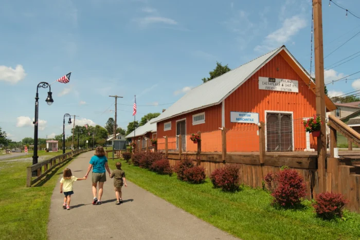 A mother and two young children walk hand-in-hand along the paved White Oak Rail Trail toward a bright orange historic freight station building.