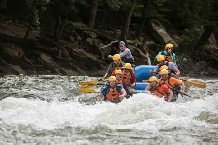 A group of whitewater rafters in yellow helmets and orange life vests navigate churning rapids on the New River.