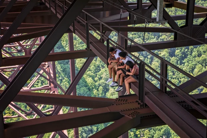Three young people sit on the catwalk of the New River Gorge bridge, gazing down at the canopy below.
