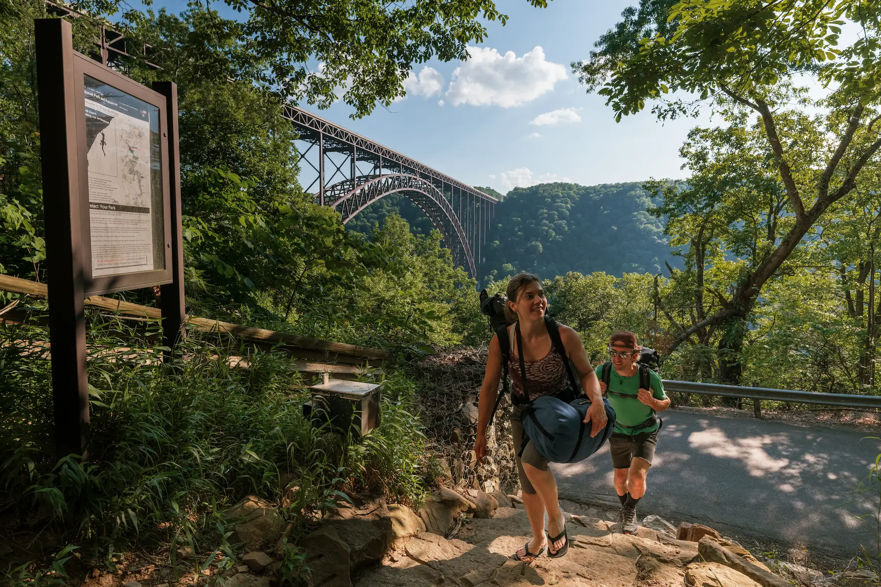 Two people climb a rocky trail through lush green trees with the iconic New River Gorge Bridge arching across the canyon in the background.
