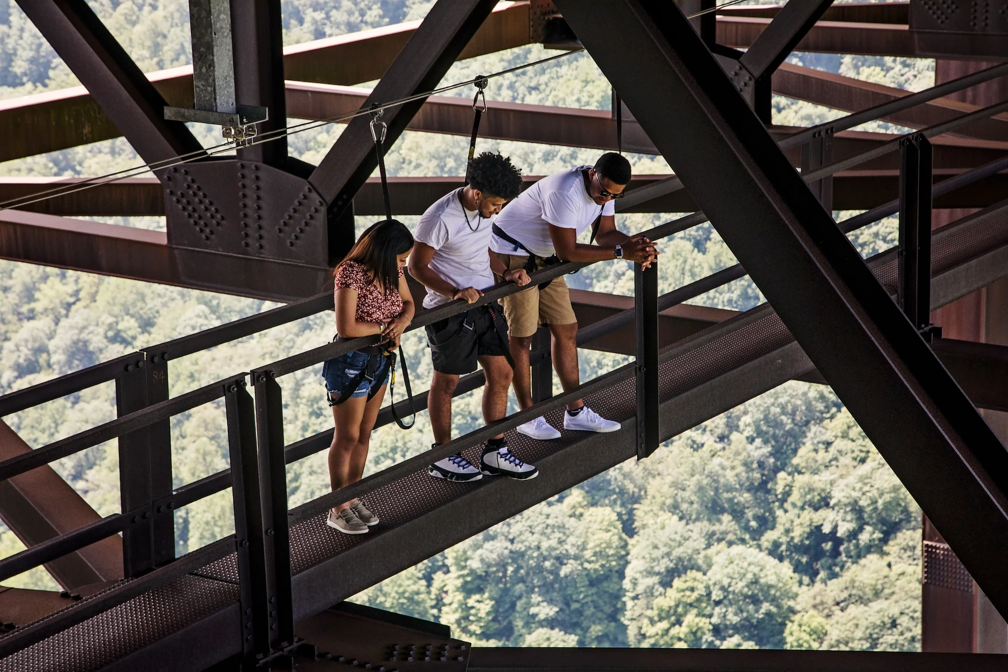 Three people wearing safety harnesses look down from a catwalk on the underside of the New River Gorge Bridge, with a tree canopy visible below.