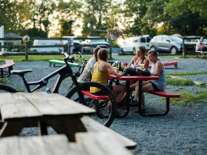 A group of women relax at a red picnic table with mountain bikes after a ride, at an outdoor patio at golden hour.