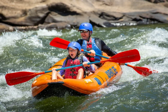 Father and daughter paddling an orange raft through whitewater rapids on the Upper New, both smiling.