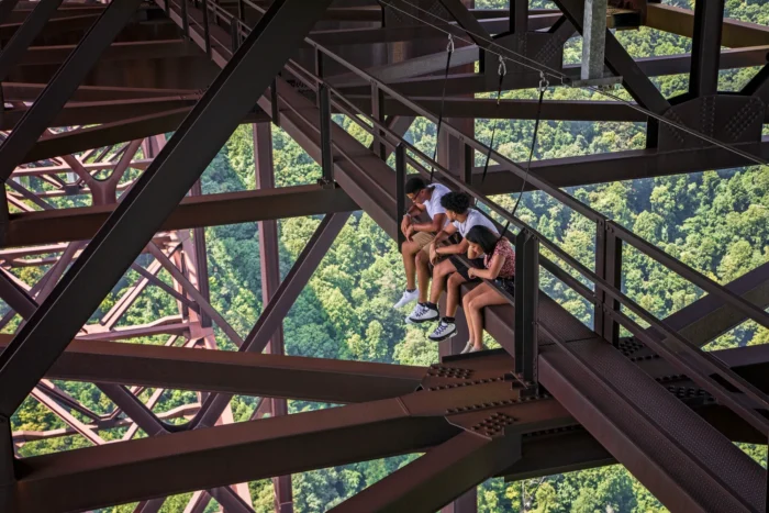 Three people sitting on a steel bridge catwalk high above a forested gorge on a Bridge Walk tour. 