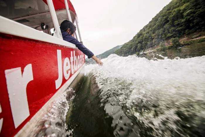 Person reaching a hand into the spray from a red New River jet boat speeding along a river.