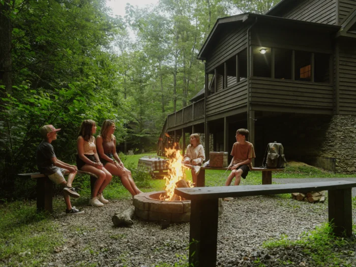 Family and friends gathered around a campfire outside a wooden cabin in the woods in the New River Gorge. 