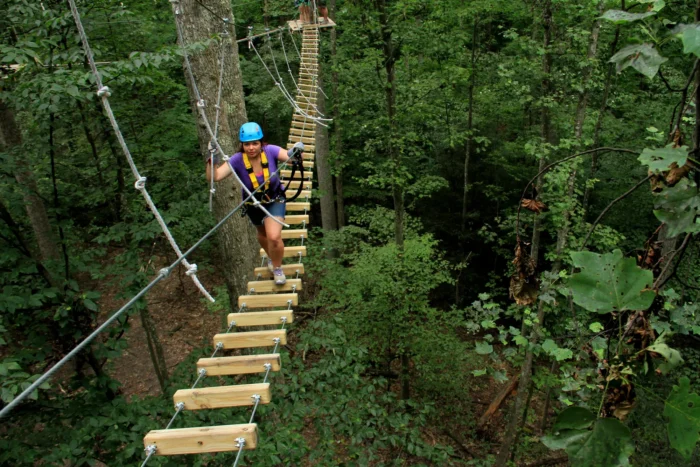Woman in helmet and harness crossing a wooden plank suspension bridge high in a forest canopy.