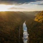 Aerial view from atop the New River Gorge Bridge on the morning of Bridge Day. The New River winds through a deep, forested gorge in autumn, with golden sunlight breaking through clouds at the horizon.