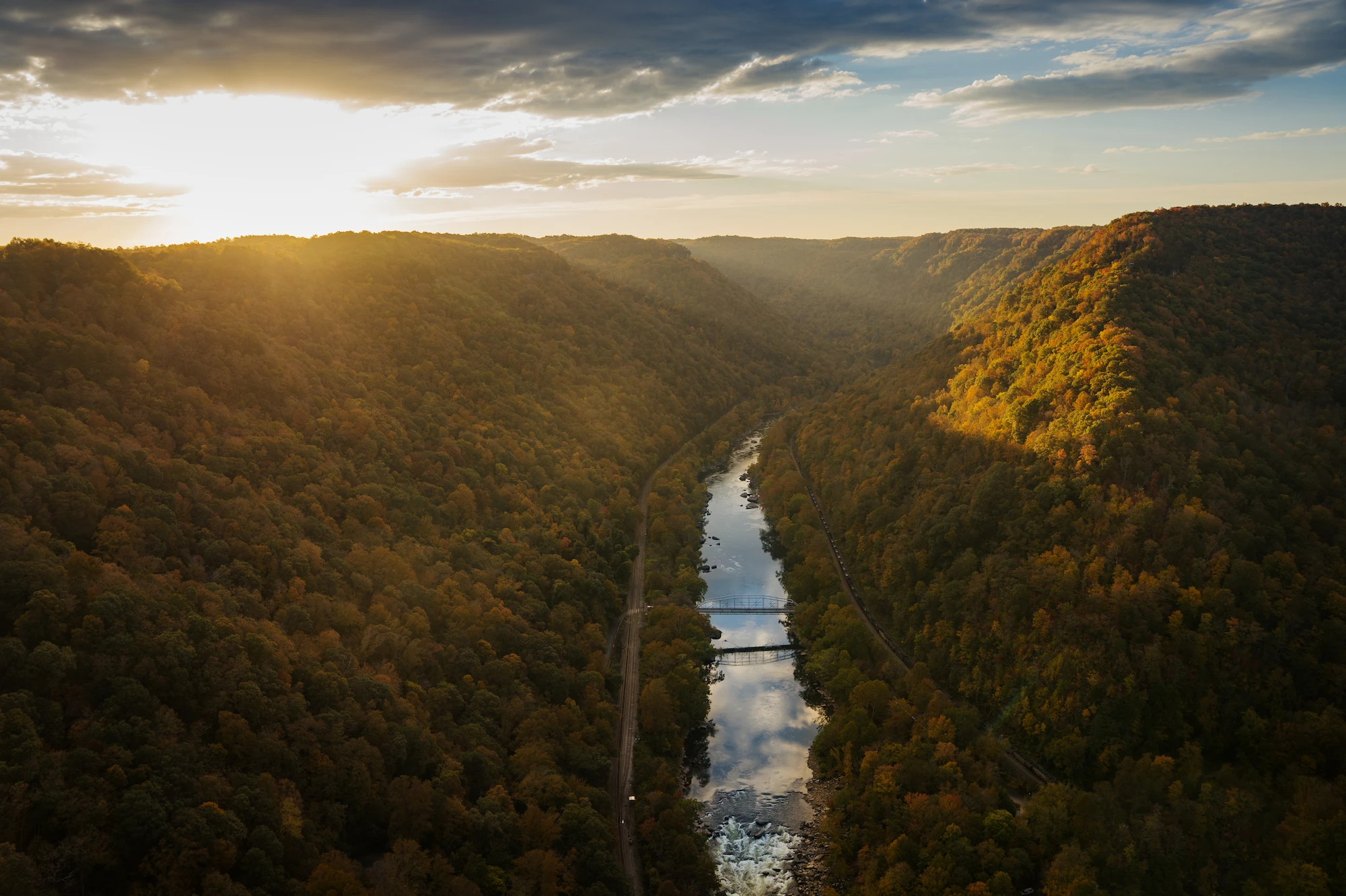 Aerial view from atop the New River Gorge Bridge on the morning of Bridge Day. The New River winds through a deep, forested gorge in autumn, with golden sunlight breaking through clouds at the horizon.